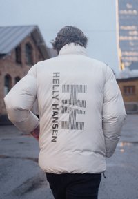 Bílá péřová bunda s výrazným logem "Helly Hansen" na zadní straně, s vertikálními pruhovanými akcenty a hladkým, lesklým povrchem.