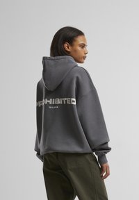 Grijze oversized hoodie met een capuchon, voorzien van vetgedrukte witte tekst "PROHIBITED" op de achterkant en een gladde, textuurstof.