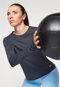 Camiseta deportiva de manga larga en tejido gris oscuro con textura acanalada, combinada con leggings azul claro, mientras sostiene una pelota medicinal negra.