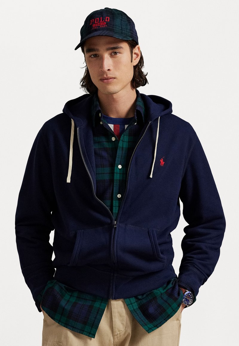 Jovem com um hoodie azul-marinho com fecho, camisa xadrez verde e azul-marinho, calças bege, boné azul-marinho com letras vermelhas e um relógio de pulso.