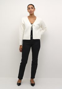 Cardigan blanc en tricot texturé avec un col en V et une fermeture à nouer, porté sur un haut noir et associé à un pantalon noir et des chaussures noires à bout pointu.
