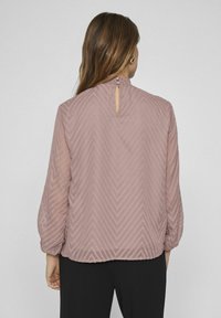 Pitkähihaista blousea mauve-värissä, jossa on teksturoitu chevron-kuvio, takana avainaukko ja pehmeä, valuva kangas. Yhdistetty mustiin housuihin.