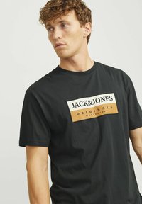 Sort kortærmet t-shirt med et rektangulært logo med hvide og brune accenter, der står "JACK & JONES ORIGINALS WORLDWIDE." Glat tekstur.