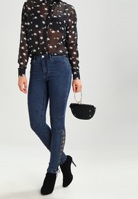 Černá průhledná blůza se vzorem hvězd spárovaná s modrými úzkými džíny s krajkovými detaily na stranách, nošená s černými podpatkovými kotníkovými botami a malou černou taškou.