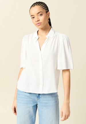 Vrouw met gevlochten haar, gekleed in een witte blouse met korte mouwen en knopen, en lichtblauwe jeans, staande tegen een effen beige achtergrond.