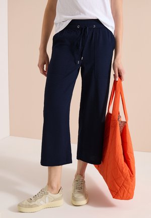 Personne porte un pantalon large bleu marine raccourci, des baskets blanches, un haut blanc et tient un grand sac cabas matelassé orange à côté.