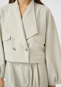 Blazer corto beige con ampi rever, doppio bottone, tasche a patta e maniche lunghe su pantaloni a vita alta plissettati abbinati.