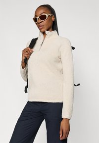 Pull en polaire beige avec un col zippé à quart, surface texturée et manches ajustées. Associé avec un pantalon sombre et des lunettes de soleil.