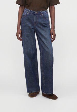 Personne portant un jean droit bleu foncé et des chaussures marron, debout devant un fond clair uni.