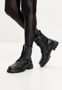 Botas de cuero negras hasta el tobillo con cordones en la parte frontal, cremallera lateral, suela texturizada y tacón grueso. Acabado suave y detalles de costura en los bordes.
