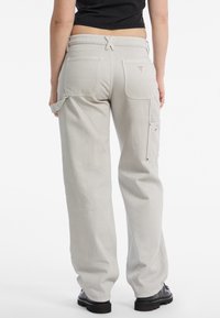 Pantaloni cargo grigi chiari realizzati in cotone, caratterizzati da una vestibilità rilassata, tasche posteriori, tasca laterale per utensili e un'etichetta con logo. Texture liscia.