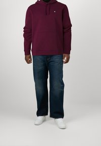Uomo che indossa un hoodie bordeaux con tasca frontale e logo, jeans blu scuro e sneakers bianche, in piedi di fronte a uno sfondo neutro.