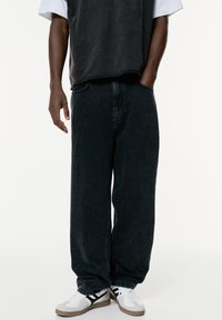 Baggy zwarte denim jeans met een vervaagde textuur, voorzien van zijzakken en een elastische zoom, gecombineerd met witte sneakers en een grijs T-shirt.