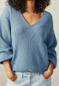 Pull tricoté bleu clair avec un col en V, des manches larges et des motifs en côtes diagonales avec de petits détails ajourés. Associé à un denim clair.