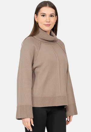 Frau mit glatten braunen Haaren trägt einen taupefarbenen Strickrollkragenpullover und schwarze Hose vor weißem Hintergrund.