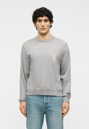 Άνδρας με σγουρά μαλλιά που φοράει ανοιχτόγριζη φούτερ με λογότυπο στο στήθος και ανοιχτό μπλε τζιν, στέκεται μπροστά σε ένα λευκό απλό φόντο.