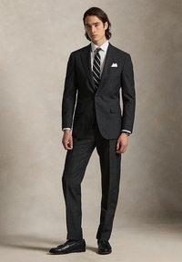 Νέος άνδρας που φοράει σκούρο κοστούμι με ρίγες, ριγέ γραβάτα, λευκό πουκάμισο, μαντηλάκι στο πέτο και μαύρα επίσημα παπούτσια, στέκεται μπροστά σε ουδέτερο φόντο.