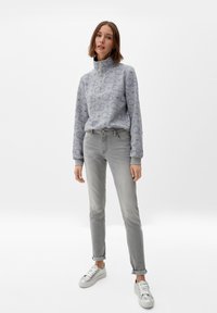 Suéter gris claro con cuello con cremallera, que presenta un sutil patrón de logotipos, combinado con jeans slim gris claro y zapatillas plateadas.
