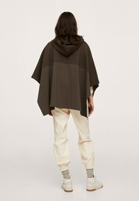Poncho à capuche marron foncé avec une coupe ample, des sections superposées et un ourlet plus long sur les côtés. Associé à un pantalon écru fuselé.