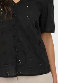 Schwarze Bluse mit kurzen Ärmeln, V-Ausschnitt, Knopfverschluss und floraler Lochstickerei mit kontrastierender innerer Fütterung.