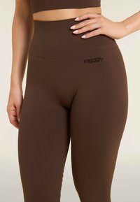 Leggings marroni a vita alta realizzati in materiale elasticizzato, con una superficie liscia e un piccolo logo nero "FREDDY" sulla vita.