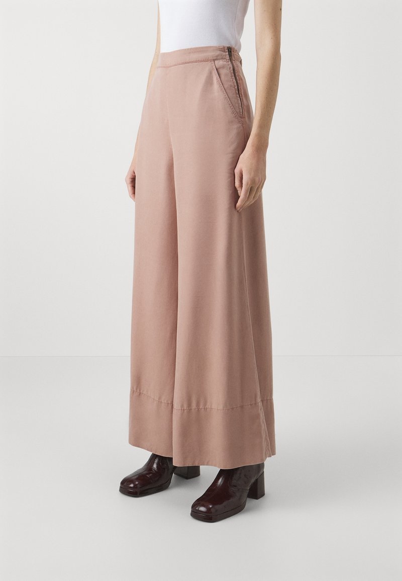 Pantalon à jambes larges en tissu doux de couleur rose clair, avec poches latérales, présentant une texture lisse et des détails de coutures discrètes, associé à des talons brun foncé.