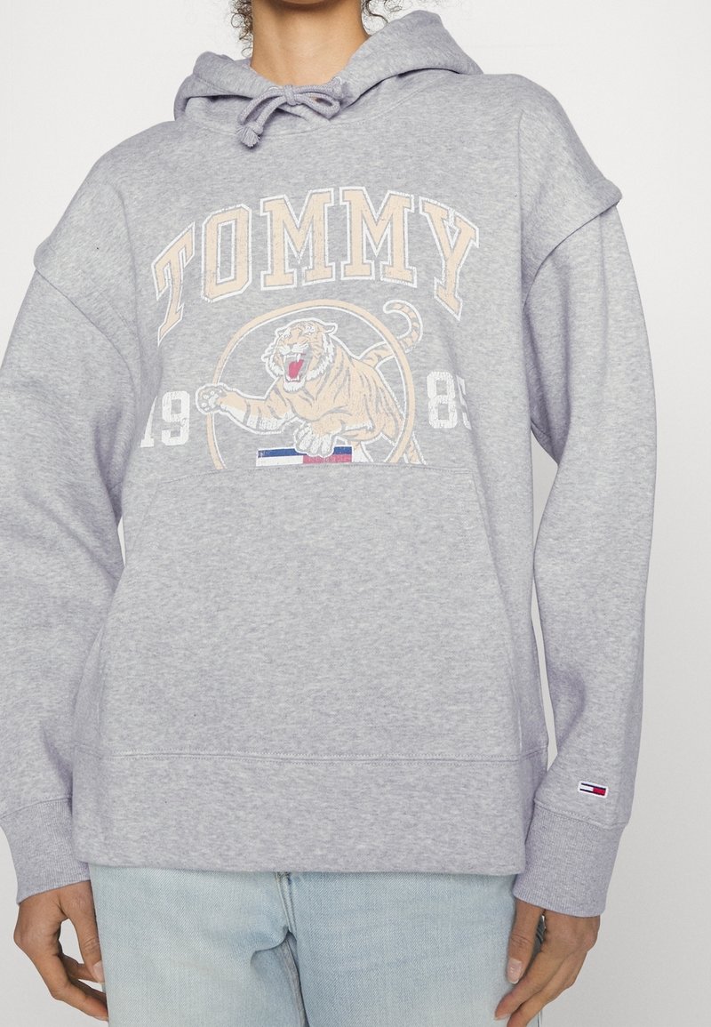 Sudadera con capucha de algodón gris con un gráfico frontal que muestra un tigre y el texto "TOMMY". Detalles incluyen un pequeño logo de bandera en la manga y capucha con cordón.