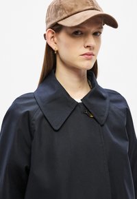 Σκούρο μπλε παλτό με μεγάλο γιακά και κλείσιμο με κουμπιά. Φορεμένο με ανοιχτό καφέ καπέλο. Λείο ύφασμα με δομημένη σιλουέτα.