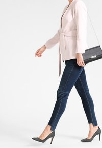 Vaaleanpunainen räätälöity bleiseri, jossa on vyötäröllä vyö, tummansiniset slim fit -farkut ja harmaat tekstuurilla varustetut korkokengät. Musta olkalaukku.