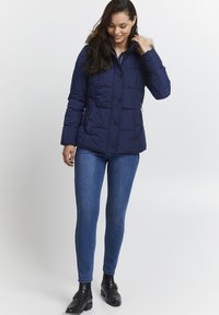 Mörkblå vadderad pufferjacka med en foderhuva i fuskpäls, tryckknappar och sidofickor, i kombination med åtsittande blå jeans och svarta ankelstövlar.