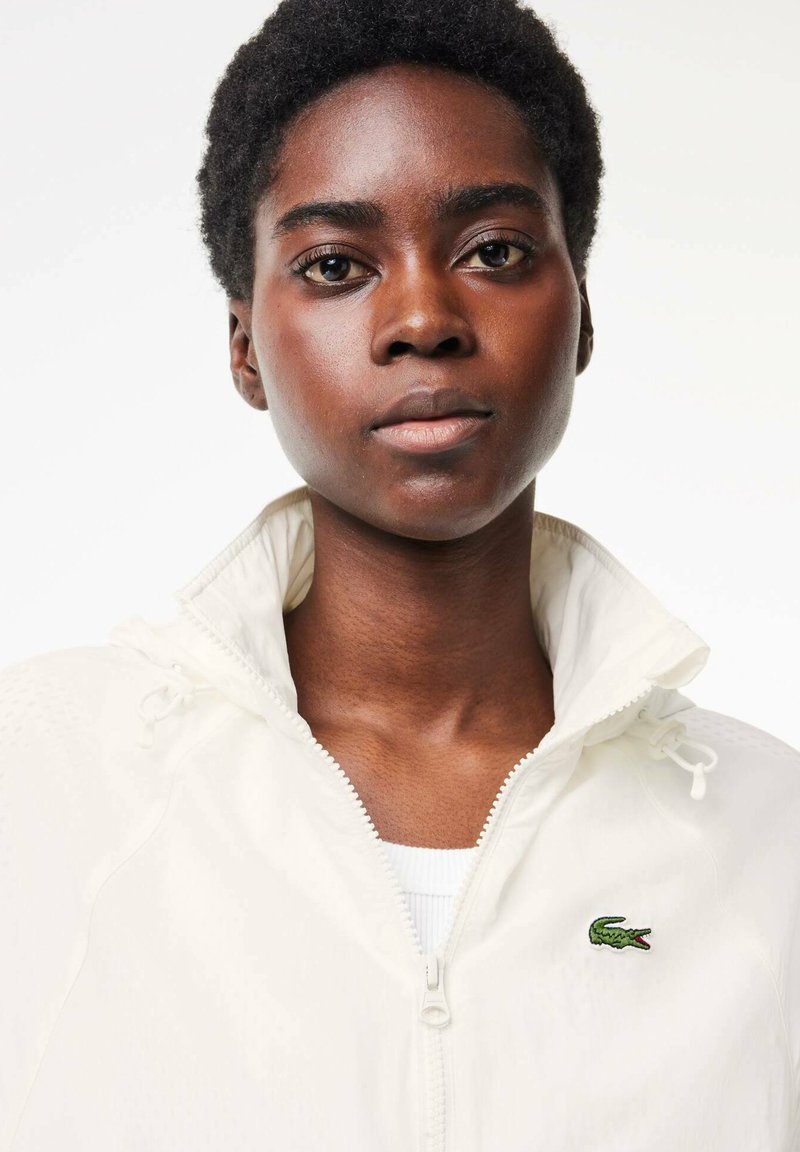 Biała kurtka zapinana na zamek z kapturem, z charakterystycznym zielonym logo krokodyla na piersi oraz teksturowanym materiałem z sznurkiem przy kołnierzu.