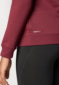 Burgunderfarget sweatshirt med ribbestrikket kant; har "WARMCELL" logo. Kombinert med svarte leggings som har en glatt tekstur og sømdetaljer.