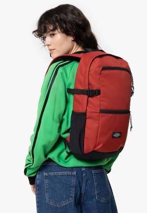 Mochila de tela roja con acentos negros, que presenta bolsillos laterales, correas ajustables y un parche con el logo. Llevada por una persona con una camiseta verde.