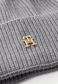 Bonnet en tricot gris avec texture côtelée, arborant un petit logo en métal doré sur le devant. Matière douce et chaude.