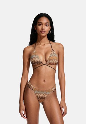 Donna che indossa un bikini marrone e multicolore con motivo a zigzag e lacci avvolgenti, in piedi davanti a uno sfondo chiaro e uniforme.