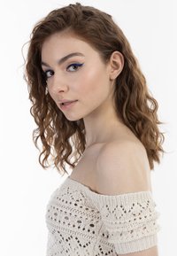 Pletený top bez ramínek v béžové barvě s texturovaným, otevřeným vzorem; modelka má vlnité, kaštanové vlasy a výrazný modrý oční make-up.