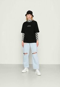 Camiseta oversized preta com texto branco, sobreposta a uma camisa de manga longa xadrez. Jeans desfiados em azul claro e sapatos de plataforma brancos. Chapéu de balde preto.