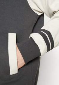 Grijze katoenen sweatshirt met crèmekleurige accenten, voorzien van een zak en geribde cuffs met zwart-witte gestreepte details. Zachte textuur.