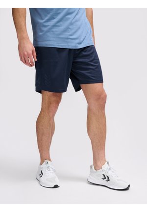 Ein Mann trägt dunkelblaue Sportshorts, ein hellblaues T-Shirt und weiße Sportschuhe und steht vor einem einfarbigen hellen Hintergrund.