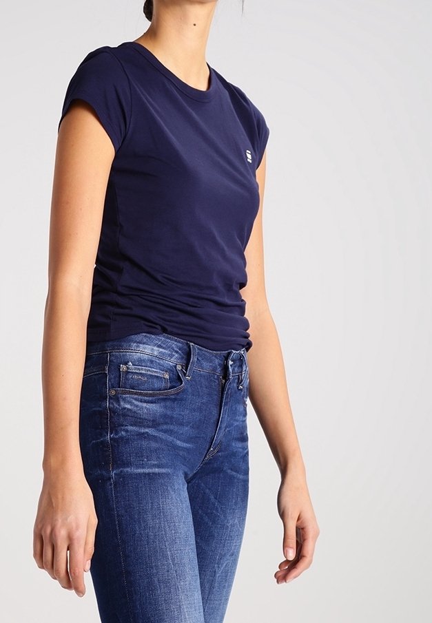 Nainen pukeutuneena istuvaan tummansiniseen lyhythihaiseen t-paitaan ja korkeavyötäröisiin sinisiin farkkuihin, seisoo yksivärisen taustan edessä.