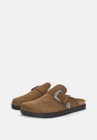 Zoccoli in suede marrone con punta chiusa e retro aperto, caratterizzati da ritagli decorativi e una fibbia argentata sul cinturino. Suola in gomma nera.