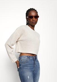 Maglione corto beige chiaro con un design a maglia testurizzata, abbinato a jeans blu strappati. Il modello indossa occhiali da sole ovali marroni.