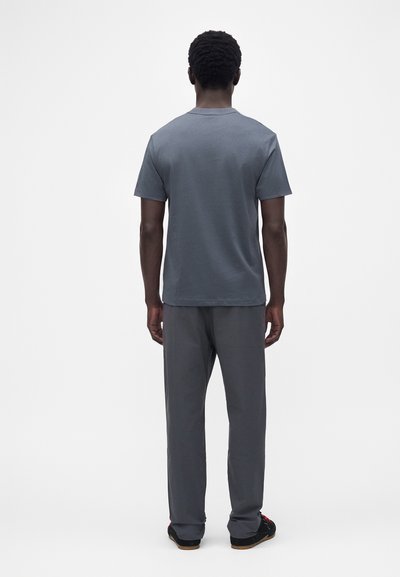 Homme debout, de dos, portant un t-shirt gris uni à manches courtes, un pantalon gris et des chaussures noires sur fond blanc.