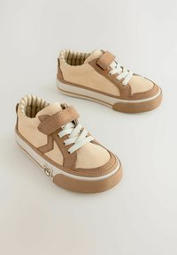 Baskets beiges avec des accents marron, lacets blancs et bande velcro. Semelle en caoutchouc texturée avec un emblème d'ours sur le côté et doublure à rayures.