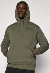 Felpa verde oliva con cappuccio regolabile, tasca a canguro frontale, scritta "PARIS" e logo Nike in nero. Tessuto morbido, polsini e orlo a costine.