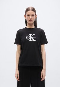 Svart bomulls-T-skjorte med en hvit logo med «Calvin Klein» og et stilisert «CK». Korte ermer og en avslappet passform. Jevn tekstur.