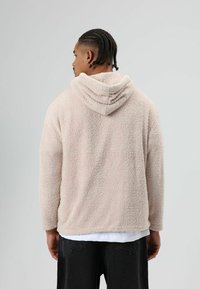 Lichtroze, pluizige fleece hoodie met een capuchon, met een gestructureerd oppervlak en lange mouwen. Draagt over een wit shirt en zwarte broek.
