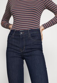 Mörkblå denimjeans med hög midja, med framfickor, kontrasterande sömmar och knäppning. Slät textur och figurnära passform.