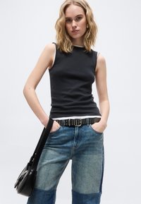 Νέα γυναίκα με κυματιστά ξανθά μαλλιά, που φοράει μαύρο αμάνικο τοπ, μαύρη ζώνη με τρουκς, τζιν με πατς και κρατάει μια μαύρη τσάντα ώμου.