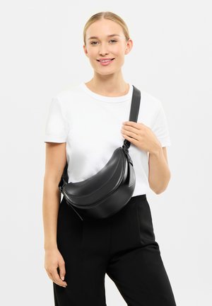 Čierna kožená crossbody taška s okrúhlym tvarom, elegantným dizajnom a nastaviteľným popruhom, nosená na ramene. Minimalistický štýl bez viditeľného kovania.